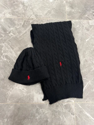 Svart kabelstickad mössa från Polo Ralph Lauren - Svart kabelstickad mössa från Polo Ralph Lauren med uppvikt kant och röd broderad logga framtill. Mjuk och varm, perfekt för kalla dagar. Snygg klassisk design som funkar till allt. Materialet är stickad ull och mössan har en normal passform.