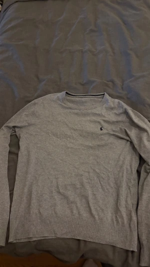 Grå stickad tröja från Ralph Lauren - Snygg grå stickad tröja från Ralph Lauren med klassisk rund hals och diskret marinblå logga på bröstet. Tröjan har ribbade muddar vid ärmslut och nederkant, och är långärmad. Perfekt för dig som gillar stilrena och enkla plagg.jag har klippt bort lappen eftersom att den kliades.skriv gärna om ni har frågor.pris kan diskuteras.
