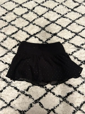 Svart kortkjol med paljetter Gina Tricot - Supersöt svart kortkjol från Gina Tricot med små glittriga paljettdetaljer. Kjolen har en lätt volangig form och bred resår i midjan för skön passform. Perfekt för dig som gillar att sticka ut lite extra med glittriga detaljer.