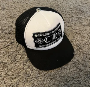 Chrome Hearts svartvit truckerkeps - Snygg truckerkeps från Chrome Hearts i svart och vitt med broderad patch framtill och metallknapp på toppen. Kepsen har mesh på sidorna och bak, samt böjd skärm. Insidan har Chrome Hearts-logga på banden. Perfekt för dig som gillar streetwear och exklusiva detaljer.