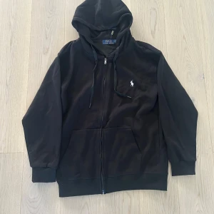 Svart zip från Polo Ralph Lauren - Klassisk svart zip hoodie från Polo Ralph Lauren med dragkedja och huva. Jackan har det ikoniska broderade logotypen på bröstet och två fickor framtill. Perfekt för en avslappnad stil. Helt ny, skriv för pris eller annat :)