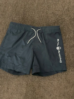 Mörkblå Sail Racing badshorts - Snygga mörkblå badshorts från Sail Racing med vit logga längs benet. Shortsen har elastisk midja med vit snörning och en bakficka med Sail Racing-märke. Perfekta för strand eller pool, stilrena och sportiga.