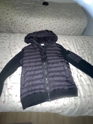 Svart pufferjacka med huva från Moncler - Svart pufferjacka från Moncler med stickade ärmar och huva. Jackan har ribbade muddar, tvåvägsdragkedja och fickor med knappstängning. Materialet är en mix av polyester och stickad textil, perfekt för kyliga dagar. Snygg, sportig look med diskret logga på insidan.Äkta