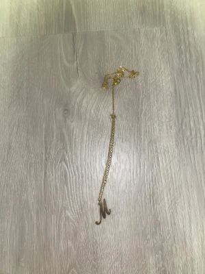 Guldigt halsband med bokstaven M - Snyggt halsband i guld med en hängande bokstav M som berlock. Kedjan är tunn och elegant, perfekt för att ge en personlig touch till din stil. Passar dig som vill ha något enkelt men ändå unikt.