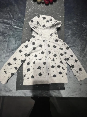 Grå hoodie med kattmönster och huva - Grå hoodie med svart kattmönster och små hjärtan över hela tröjan. Tröjan har dragkedja framtill, långa ärmar och en mysig huva. Perfekt för dig som gillar gulliga detaljer och vill ha en chill stil.