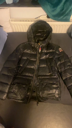 Svart parajumper jacka - Svart dunjacka från Parajumpers, modell Sheen, Jackan har en glansig finish, huva, två stora fickor framtill och dragkedja. Klassisk patch på ärmen och coola detaljer invändigt. Perfekt för kalla dagar