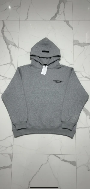 Grå Essentials Fear of God hoodie - En klassisk grå hoodie som passar perfekt för vardag och mysiga stunder. Egenskaper: 	•	Mjuk och bekväm: Gjord för maximal komfort. 	•	Klassisk design: Enkel, stilren och lätt att matcha med allt. 	•	Justerbar huva: Perfekt för alla väder. 	•	Ribbad mudd: Håller formen och sitter snyggt.  Skick: Helt ny Färg: Grå Storlek: L