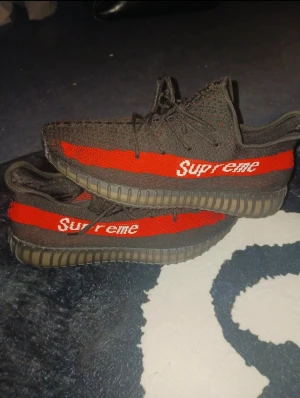 Adidas Yeezy Boost Supreme svart/röd - Säljer ett par Adidas Yeezy Boost sneakers med Supreme-tryck. Skorna är svarta med en röd stripe och vit Supreme-text på sidan. De har stickad ovandel, snörning och en genomskinlig, räfflad platt sula. Perfekt för dig som gillar streetwear och unika samarbeten.