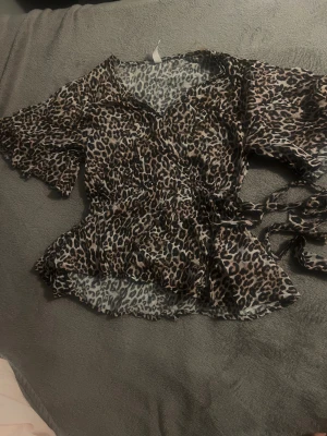 Leopardmönstrad omlottblus med volang - Säljer en snygg leopardmönstrad omlottblus med volangdetaljer och knyt i midjan. Blusen har korta ärmar och v-ringad hals. Materialet är lätt och luftigt, perfekt för att sticka ut med ett djuriskt mönster. 