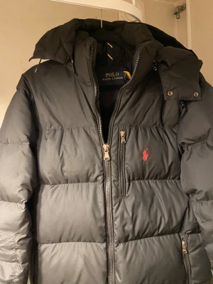 Svart  jacka från Polo Ralph Lauren - Svart pufferjacka från Polo Ralph Lauren med röd broderad logga på bröstet. Jackan har hög krage, avtagbar huva och flera dragkedjeförsedda fickor. Fylld med dun och har en stilren, quiltad design. Perfekt för kalla dagar och snygg till streetwear.