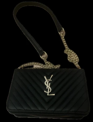 YSL väska - Snygg svart axelväska från YSL med guldiga kedjedetaljer och ikonisk YSL-logga framtill. Väskan är quiltad i ett V-format mönster och har en stilren, rektangulär form. Tillverkad i skinn med både kedja och läder på axelremmen för extra komfort.