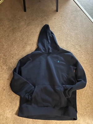 Marinblå hoodie från Polo Ralph Lauren - Säljer en marinblå hoodie från Polo Ralph Lauren i storlek M. Tröjan har en klassisk huva, stor magficka och det ikoniska Polo-logot broderat i ljusblått på bröstet. Perfekt för chill dagar och snygg till jeans eller joggers.