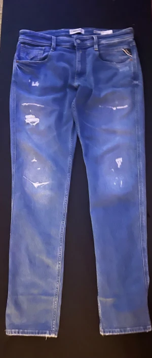 Blå Replay Anbass jeans med slitningar - Snygga blå jeans från Replay, modell Anbass. Jeansen har en slim passform och är tillverkade i klassiskt jeansmaterial. De har coola slitningar och detaljer på benen som ger en trendig look. Perfekt för dig som gillar streetstyle och vill sticka ut.