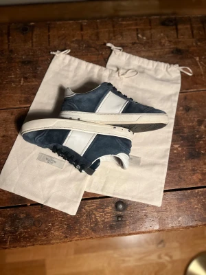 Valentino Garavani blå sneakers mocka - Snygga sneakers från Valentino Garavani i blå mocka och vitt skinn. Klassisk låg modell med svarta snören, vita detaljer och coola nitar på sulan. Sulan har ett unikt mönster undertill. Perfekta för dig som vill ha en stilren men ändå lyxig look. Köptes här på plick i börkan av året. Storlek 38 men passar även 39. Skick mellan 7/10 och 8/10. Ena skon saknar 3 nitar, annars är dom i bra skick, skor och dustbags hänger med. Priset går att diskuteras! Tveka inte att skriva om du har någon fråga!🤗🙌