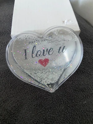 Hjärtfotohållare med glitter - Säljer en hjärtformad fotohållare med texten 'I love u' och ett rött hjärta i mitten. Fylld med flytande silverglitter som rör sig när du vänder på den. Perfekt som present eller dekoration på skrivbordet. Helt ny med förpackning.