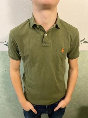 Olivgrön pikétröja från Polo Ralph Lauren - Snygg olivgrön pikétröja från Polo Ralph Lauren med klassisk krage och två knappar framtill. Tröjan har korta ärmar och den ikoniska orangea Polo-loggan broderad på bröstet. Tillverkad i mjuk bomull som är skön mot huden.