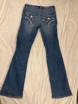 Blå bootcut jeans med paljettdetaljer - Snygga blå jeans med bootcut passform och coola paljettdetaljer på bakfickorna. Jeansen har klassisk femficksdesign, låg midja och dekorativa sömmar. Perfekta för dig som gillar lite extra detaljer på dina jeans.