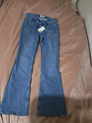 Blå bootcut jeans från 157 - Bootcut jeans från 157 i klassisk blå med låg midja. Modellen heter Low Boot Patch och har fickor, bälteshällor och knappstängning. Jeansen är tillverkade i jeansmaterial och har en snygg utsvängd passform nedtill. De är klippta se bild 97cm