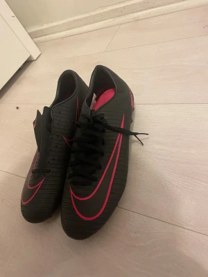 Nike Mercurial svarta fotbollsskor - Säljer ett par Nike Mercurial fotbollsskor i svart med rosa detaljer och logga. Skorna har snörning, rund tå och platt sula med dobbar för gräsplan. Yttermaterialet är syntet med diskret randig struktur och insidan är rosa. Perfekt för dig som vill sticka ut på planen. Säljer pågrund av att de är för små. Aldrig använd. Står att det är storlek 40 men skulle säga passar 38/39.