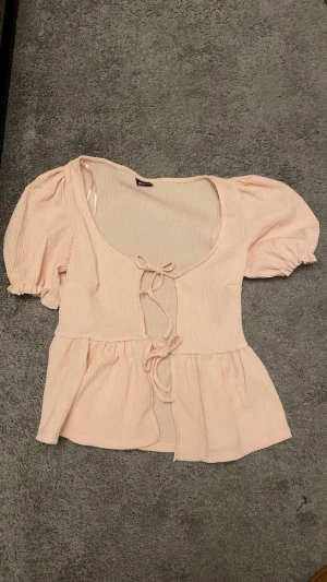 Ljusrosa blus med puffärm och knyt - Supersöt ljusrosa blus med puffiga korta ärmar och två knyt framtill. Blusen har en lätt rynkad struktur och volang nedtill som ger en romantisk vibe. Perfekt för dig som gillar en feminin och trendig look.