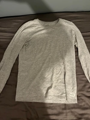 Grå långärmad tröja från 157 - En enkel och stilren grå långärmad tröja från 157. Tröjan har rund halsringning och är gjord i mjuk bomullsmix som är skön mot huden. Perfekt att ha som basplagg och funkar till många olika outfits. Diskret logga nertill på framsidan.