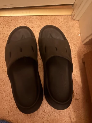 Svarta sandaler från Hoka - Svarta sandaler från Hoka med bred rem över foten och ventilerande hål. Sulan är tjock och har tydligt mönster för extra grepp. Tillverkade i syntetmaterial och har en platt sula med rund tå. Perfekt för chill dagar eller efter träning.