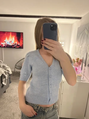 Ljusblå stickad kofta med kort ärm - Supersöt ljusblå stickad kofta med v-ringning och korta puffärmar. Koftan har ribbade muddar och knappar framtill, perfekt att styla med jeans för en chill look. Mjuk och skön, passar både till vår och sommar.