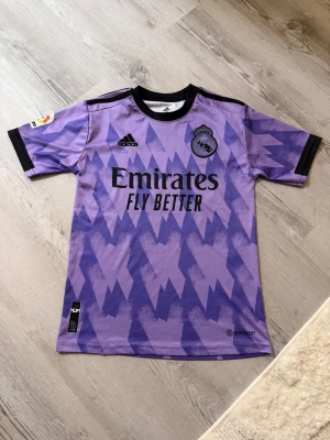 Real Madrid Vinicius Jr. lila matchtröja - Säljer en Real Madrid matchtröja från Adidas i lila med svart detaljer och grafiskt mönster. Kortärmad med La Liga-märke på ärmen, klubbmärke på bröstet och tryck med Vinicius Jr. 20 på ryggen. Materialet är lätt polyester, perfekt för fotboll.Det står att de är storlek 176 men det är 164