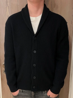 Svart ribbad kofta från Massimo Dutti - Snygg svart kofta från Massimo Dutti i ribbstickad ull. Modellen har lång ärm, bred krage och knäppning med fem svarta knappar framtill. Perfekt för dig som gillar stilrena och klassiska plagg med lite extra edge.