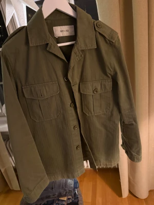 Olivgrön overshirt från Whyred - Snygg olivgrön overshirt från Whyred med klassisk krage, axelklaffar och två stora bröstfickor med knappar. Jackan har raka linjer och knäppning framtill, perfekt för lager-på-lager. Materialet är bomull och modellen är långärmad.