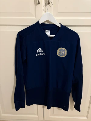 Marinblå GIF Sundsvall fotbollströja - Säljer en marinblå långärmad fotbollströja från Adidas med GIF Sundsvall-emblem på bröstet och vita Adidas- och Stadium-loggor. Tröjan har V-ringad hals och är tillverkad i ett lätt, funktionellt material som passar perfekt för träning eller match.