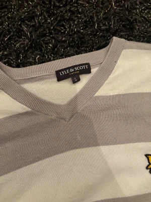 Randig Lyle & Scott tröja i grått och vitt - Snygg stickad tröja från Lyle & Scott i storlek L med breda grå och vita ränder. V-ringad hals och klassisk gul logga på bröstet. Långärmad med ribbade muddar och mjukt material som känns skönt mot huden. Perfekt för dig som gillar stilren och chill look.