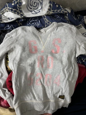 Grå sweatshirt från G-Star RAW - Grå sweatshirt från G-Star RAW med tryck i rosa på framsidan och ribbade muddar vid ärmslut och nederkant. Klassisk rund hals och mjukt bomullsmaterial. Perfekt för chill dagar och streetwear-stil.