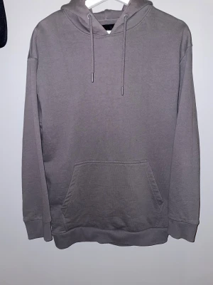 Grå hoodie med ficka och huva - En stilren grå hoodie med klassisk känguruficka framtill och justerbar snörning i huvan. Tröjan har långärmade ärmar och ribbade muddar vid ärmslut och nederkant. Perfekt för chill dagar och enkel att matcha med jeans eller joggers. Köpt från Lager 157 och har knappt använts.