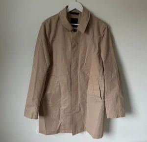 Beige trenchcoat från J.Lindeberg, stl 48 - Stilren beige trenchcoat från J.Lindeberg i mjuk twill. Jackan har klassisk krage, dold knäppning framtill och två sneda fickor. Tillverkad i 100% polyester och har knappar vid ärmslut. Perfekt för dig som gillar minimalistisk och clean look. 5K12