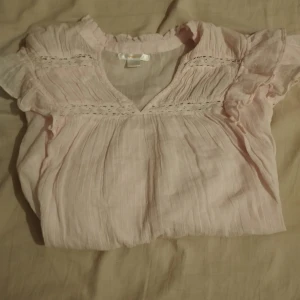 Ljusrosa blus från Gina Tricot - Supersöt ljusrosa blus från Gina Tricot med volangdetaljer vid axlarna och v-ringad hals. Blusen har långärmade ärmar med spetsinslag och ett luftigt, skirt tyg i bomull. Perfekt för dig som gillar romantisk och feminin stil.