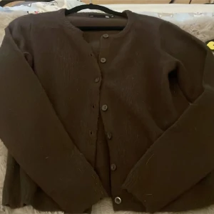 Brun kofta från 157 med knappar - Mysig brun kofta från 157 i storlek XS. Koftan har rund hals, långa ärmar och stängs med fem knappar framtill. Materialet är stickat och känns mjukt mot huden, perfekt för kyliga dagar. Enkel och stilren design som funkar till många olika outfits.