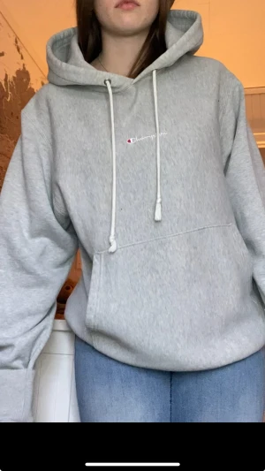 Grå hoodie från Champion med huva - Säljer en ljusgrå hoodie från Champion med broderad logga på bröstet. Tröjan har huva med snörning, magficka och ribbade muddar. Oversized passform och mjukt material,s i kill-storle, defekt på snöret