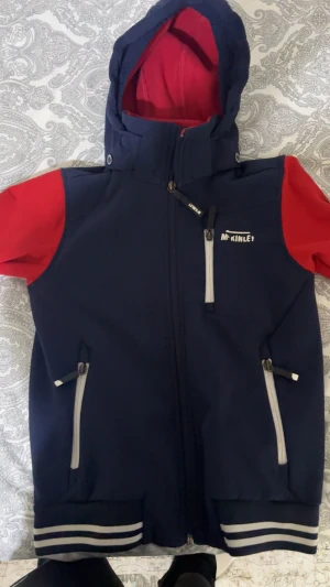 Blå och röd vindjacka från McKinley - Snygg vindjacka från McKinley i marinblått med röda ärmar och huva. Jackan har dragkedja framtill, tre fickor med dragkedja och coola vita ränder på mudden. Insidan är fodrad med röd fleece för extra värme. Perfekt för utomhusaktiviteter!