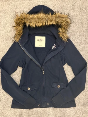 Hollister kofta med päls - Snygg mörkblå jacka från Hollister med huva och fluffig päls. Jackan har dragkedja, knappar på fickorna och broderad logga på bröstet. Perfekt för kyliga dagar till vintern eller hösten💕