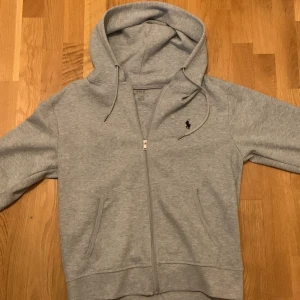 Grå hoodie från Polo Ralph Lauren - Grå hoodie från Polo Ralph Lauren med dragkedja och huva med snörning. Helt oanvänd. Klassisk svart logga broderad på bröstet. Mjuk bomullsmix, två fickor fram och ribbade muddar. Perfekt för chill dagar och enkel att matcha med jeans eller joggers.