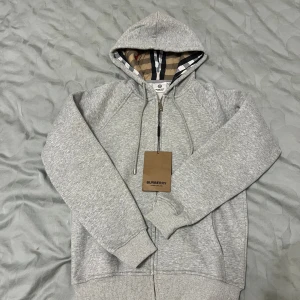 Grå hoodie från Burberry med dragkedja - Snygg grå hoodie från Burberry med klassiskt rutigt foder i huvan. Tröjan har snörning vid huvan, ribbade muddar och en praktisk dragkedja framtill. Perfekt för dig som gillar stilrena och exklusiva plagg med en twist. Storlek M men passar som S