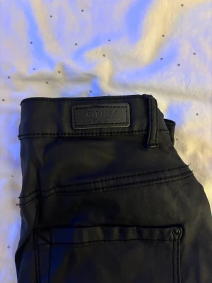 Svarta coated jeans från ONLY, S/30 - Säljer ett par svarta coated jeans från ONLY i storlek S/30. Byxorna har en snygg glansig finish som ger en cool look och klassiska bakfickor. Passformen är slim och materialet känns mjukt men ändå stadigt. Perfekta till sneakers eller boots.