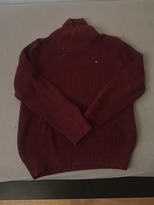 Vinröd stickad tröja Tommy Hilfiger - Säljer en vinröd stickad tröja från Tommy Hilfiger med snygg krage och knappdetalj. Tröjan har ribbad struktur, lång ärm och liten broderad logga på bröstet. Perfekt för dig som vill ha en klassisk men ändå modern look.