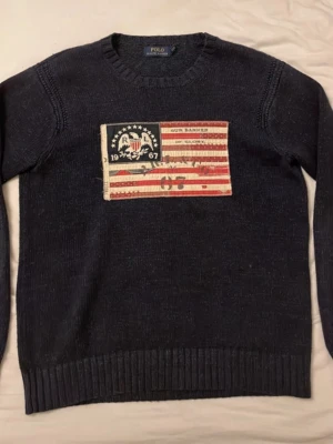 Mörkblå stickad tröja Polo Ralph Lauren - Säljer en mörkblå stickad tröja från Polo Ralph Lauren med en stor vintageinspirerad patch framtill i amerikansk flaggstil och texten 'Our Banner of Glory'. Tröjan har rund hals, långa ärmar och ribbade muddar. Perfekt för dig som gillar klassisk college-stil.