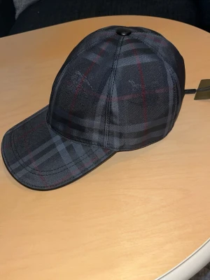 Rutig Burberry keps i mörka toner - Säljer en snygg rutig keps från Burberry i mörka färger med inslag av rött och grått. . Helt ny oanvänd. Tar emot bud.
