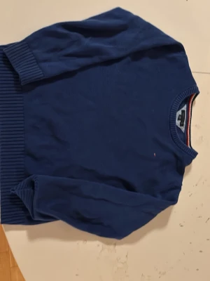 Marinblå stickad tröja Tommy Hilfiger - Säljer en marinblå stickad tröja från Tommy Hilfiger i storlek XL. Tröjan har ribbade muddar vid ärmslut och nederkant samt en klassisk rund halsringning med röd, vit och blå rand. Liten broderad logga på bröstet. Perfekt för dig som gillar stilren design.