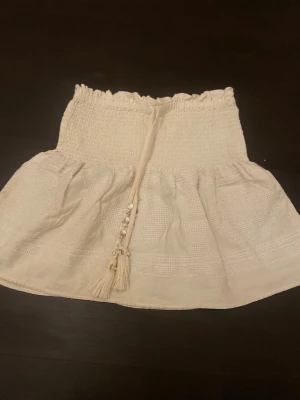 Vit kortkjol med snörning och tofsar - Supersöt vit kortkjol med smock i midjan och dekorativ snörning framtill med pärlor och tofsar. Kjolen har en lätt volang och är gjord i ett luftigt bomullstyg med diskret struktur. Perfekt för varma dagar och ger en bohemisk vibe.