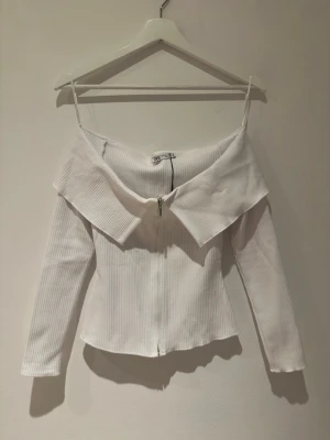 Vit ribbad offshoulder topp från Zara - Säljer en vit ribbad topp från Zara i storlek S. Toppen har offshoulder-design med smala axelband, lång ärm och en snygg dragkedja framtill. Materialet är stretchigt och ger en slim passform. Perfekt för dig som gillar trendiga detaljer och stilren look. (HELT NY MED KVITTO PÅ)