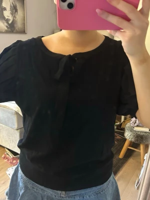 Svart blus med knyt och puffärm - Säljer en svart blus med rund halsringning och knyt fram. Blusen har korta puffärmar med veckade detaljer som ger en snygg siluett. Materialet känns mjukt och lätt, perfekt till jeans eller kjol.